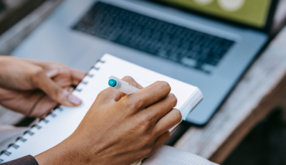 Henkilö suunnittelee kesäsesongin hyödyntämistä kirjoittamalla muistikirjaan ja käyttämällä kannettavaa tietokonetta ulkona.
